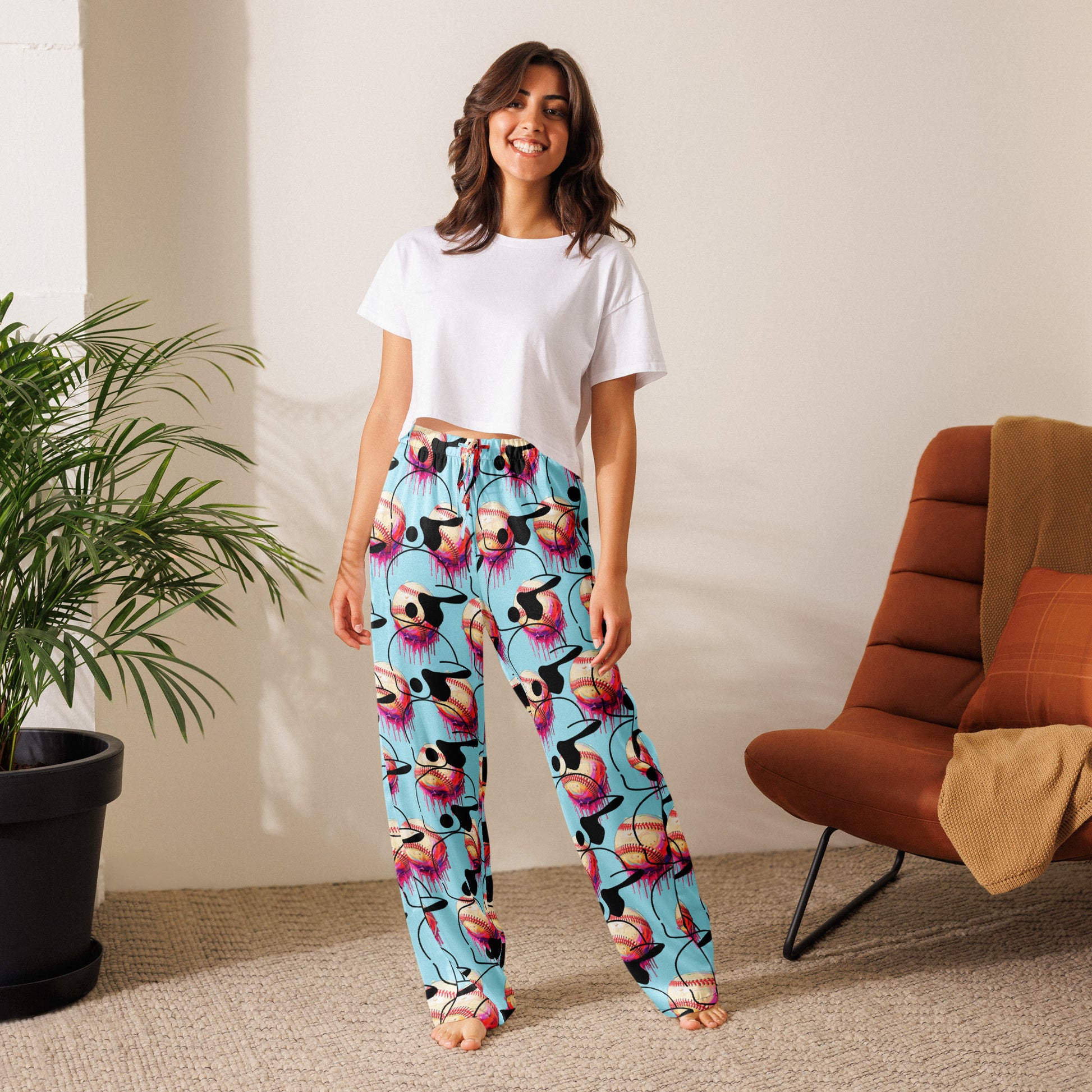 A woman in a relaxed fit white t-shirt and Steams Baseball Women’s pajama pants smiles barefoot indoors. Her loungewear suits the cozy setting, with a plant nearby and a brown cushioned chair on the carpeted floor.